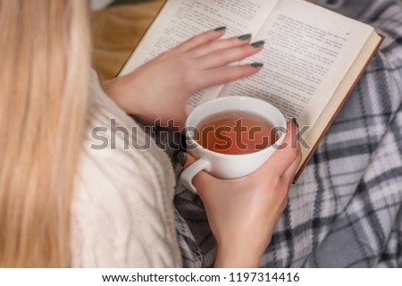 Woman holds hot cup of tea and reading book in bed. Girl wears woolen sweater and covered legs with retro blanket. Winter and cold weather concept. Close up, selective focus