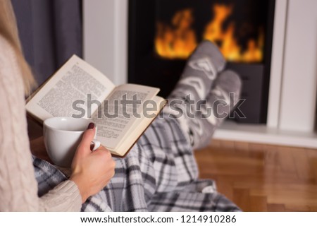 Girl in front of the fireplace reading book and warming feet on fire and legs are covered with blanket, in hand holds cup of hot tea. Winter and cold weather concept at home. Close up, selective focus