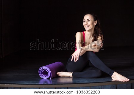 Rest after workout in the gym. Sport beautiful smiling woman in sportswear with a yoga mat sitting and relaxing after yoga or fitness.