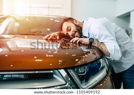 Happy handsome bearded man buying a car in dealership, guy hugging hood of new car