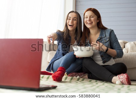 Two girl friends sitting on the floor near the couch in front of them there is a computer on which they watch a movie and eat popcorn. Funny friends relaxing together.