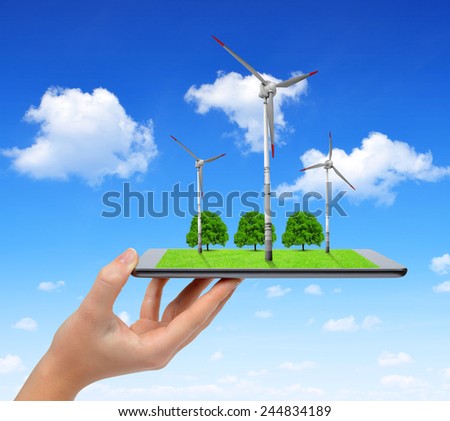 Hand holding digital tablet with a wind turbines and trees . Green energy concept.
