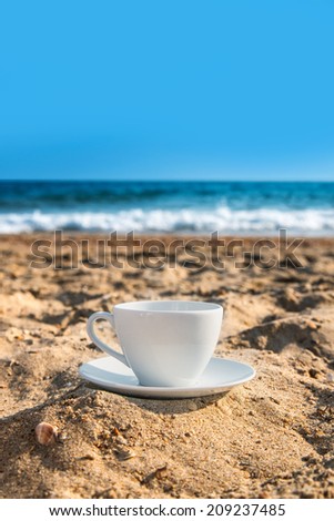 white cup with tea or coffee on sand beach front of sea, close up