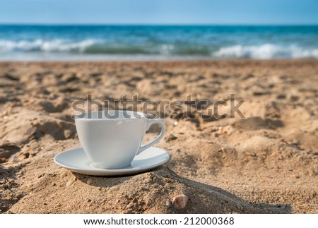 white cup with tea or coffee on sand beach front of sea, close up