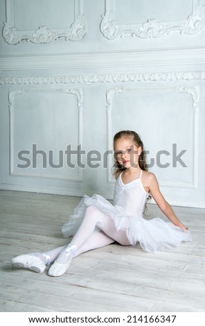 A little adorable young ballerina in white ballet dress tutu is sitting on floor in the interior studio