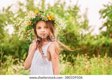 Cute smiling little girl with flower wreath on the meadow at the farm. Portrait of adorable small kid outdoors. Midsummer. Earth Day