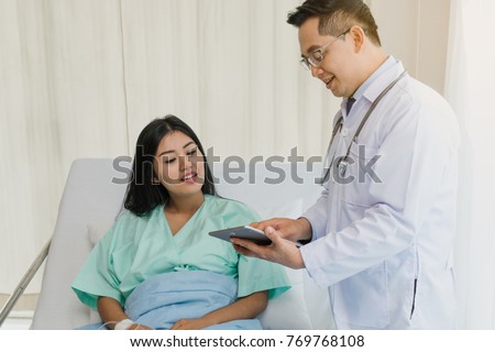 cheerful Asian doctor talking with young woman patient for monitoring and check up after surgery. doctor giving advice in the touchscreen to women to take care the lesion after out of hospital.