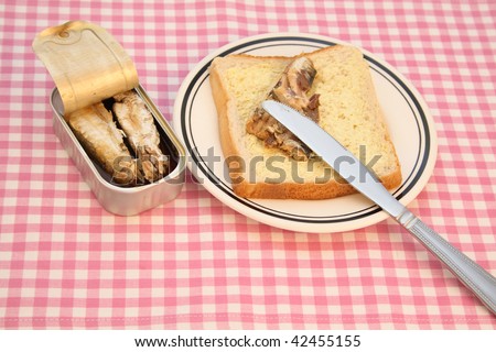 Open sardine tin and a slice of bread on a checkered table cloth.