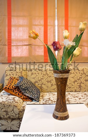 a corner of parlor shows flowers in vase on a white table ,fabric sofa , pillows and drapes