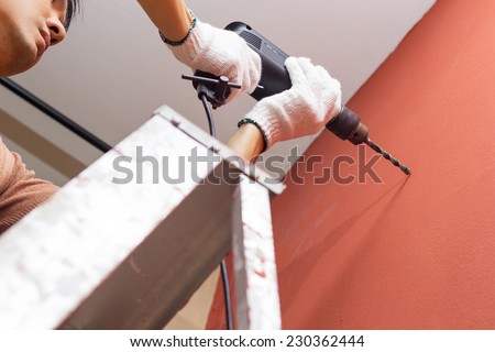 asian handsome man with electric drill on ladder making hole in brown wall / repair, renovation , building and home interior concept