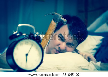 sleeping asian young male disturbed by alarm clock early morning on bed & holding hammer in hand