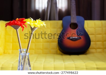 red & yellow flowers in glass vase on sunburst acoustic guitar & yellow fabric armchair background
