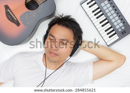 Sleeping asian man lying on white bed while listening music through headphone & guitar, piano