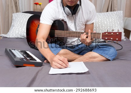 asian male music composer, musician writing song & playing guitar on bed in bedroom