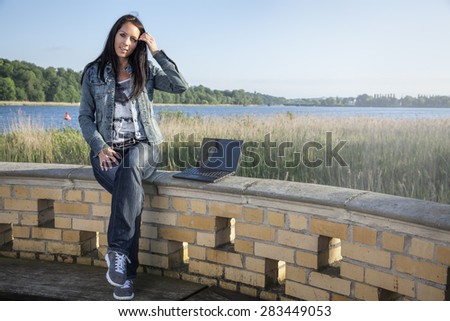 Beautiful woman is sitting on a wall near by a lake with a notebook