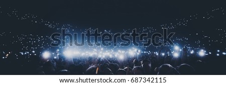 Group of people holding cigarette lighters and mobile phones at a concert \
crowd of people silhouettes with their hands up. Dark background, smoke, spotlights. Bright lights