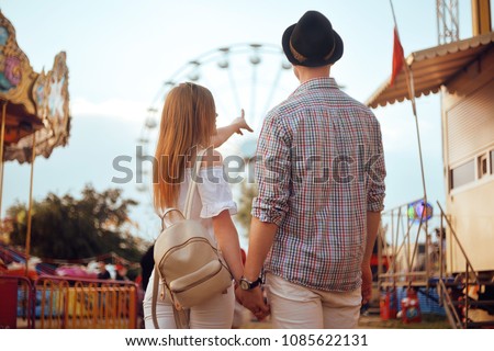 Beautiful, young couple having fun at an amusement park. Couple Dating Relaxation Love Theme Park Concept. Couple posing together on the background of a ferris wheel. Tourists have fun, smile.