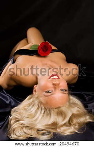 Beautiful blonde young woman with red rose laying on black satin sheets. Shot upside down
