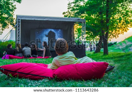 Boy back laying cozy on pillow i green grass and watching film in open cinema in public green park.Perfect spending weekend time in open air.