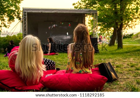 Young woman and girl laying cozy on pillow i green grass and watching film in open cinema in public green park.Back of twofemale in nature enjoying movie on big screen