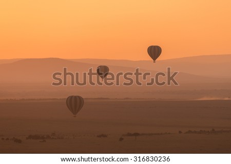 Three hot air balloons fly over the low hills of the African savannah. The aerial perspective makes the hills look different shades of purple, and the sky is bright orange in the pre-dawn light.