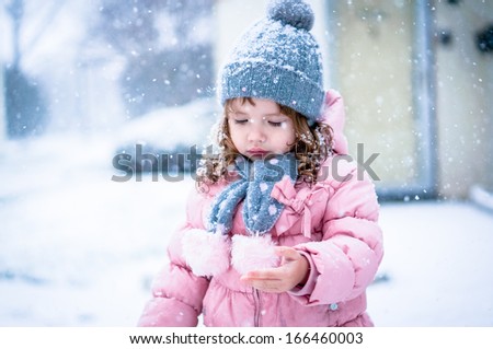 Cute baby girl in pink jacket and grey hat enjoying first snow blowing