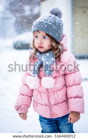 Cute baby girl in pink jacket and grey hat enjoying first snow blowing