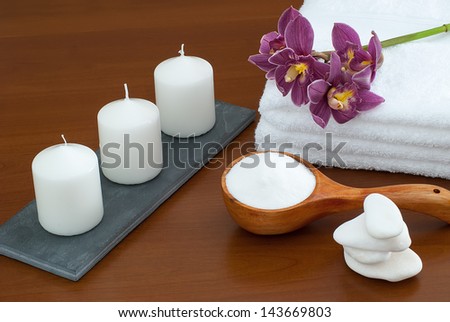 Spa still life with sea salt scrub exfoliation, orchid flower and stack of white towels in the bathroom with candles and hot stones foreground