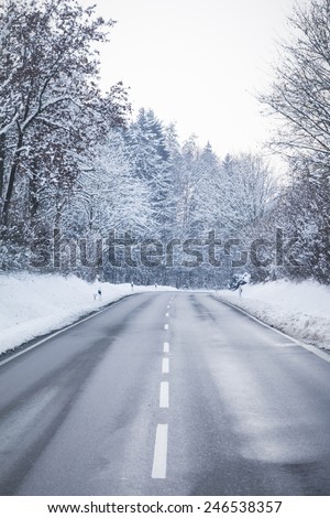 Cold winter road with wonderful white snow covered forest trees. The country street is wet and has a curcve in cold colors.