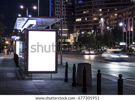 Blank poster on bus stop at night. Copy space. In background out of focus street, cars, buildings.