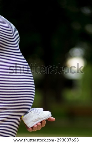 woman holding little baby shoes in front of the pregnant belly. Close up picture of pregnant woman holding a baby shoe