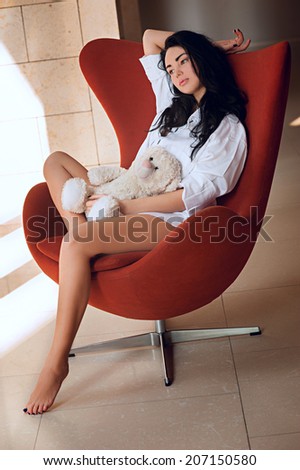 young girl with a toy in chair,girl on a red chair in a white shirt standing near window