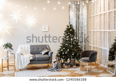 Christmas decor. Bright interior with a large Christmas tree, a gray cozy sofa and large bright garlands on the white walls.