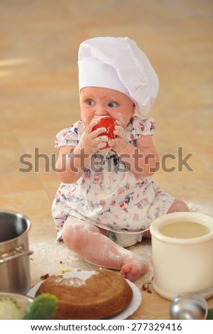 A little cute baby-girl with cooking hoods on her head is sitting on the kitchen floor, she is heavily soiled with wheat flour - she\'s playing cook and bites her teeth into a big red tomato