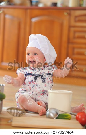 A little cute ten-months-baby-girl with cooking hoods on her head is sitting on the kitchen floor, she is heavily soiled with wheat flour - she's playing cook and she is very happy about it