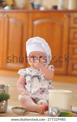 A little cute baby-girl with cooking hoods on her head is sitting on the kitchen floor, she is heavily soiled with wheat flour - she's playing cook