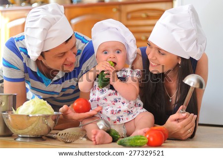 Young family with cooking hoods on their heads: a little cute baby-girl is sitting on the kitchen floor, she is eating cucumber - her parents are lying next to her smiling and playing cooks