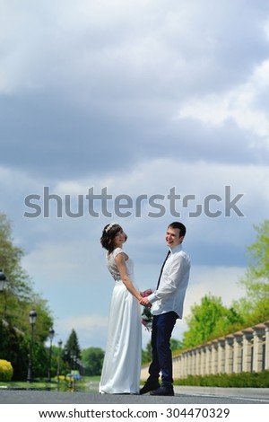 Very young and beautiful bride and groom turned smiling to the photographer - a full-length portrait - and on a background of white, blue and purple sky before the storm