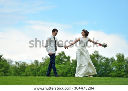 The bride pulls the hand of his beloved fiance and beckons to follow her - under their feet is luscious green grass and over their heads - bright blue sky. They are happy