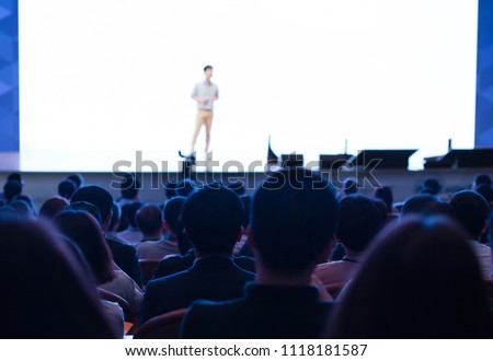 White Background Wall Screen for Presentation. Presenter Presenting on Stage at Conference Auditorium Hall Exhibition. Lecture Speaker. Blurred De-focused Unidentifiable Presenter and Audience.