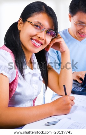 Female scholar smiling to the camera while the male scholar on the background typing on his laptop