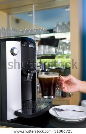 A hand is holding a glass of coffee that is ready to be served from the coffee maker