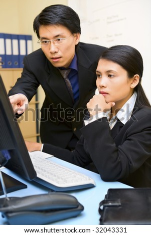 A business man is pointing at the computer screen which his partner works on.