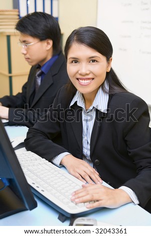 A woman employee is smiling in her office, while the working man behind her is out of focus.