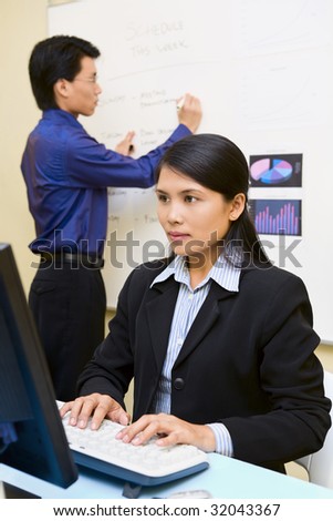 An employee woman is working on her computer while the man in background is busy writing on the whiteboard.