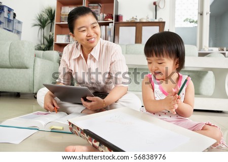 LIttle Chinese girl playing with drawing paper with her mother or nanny on background