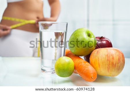 Fruit and water on table with female body on sport attire at background measuring h er stomach. shot for diet concept