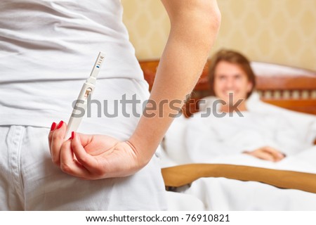 A lady is hiding her pregnancy test in front of her smiling spouse on bed