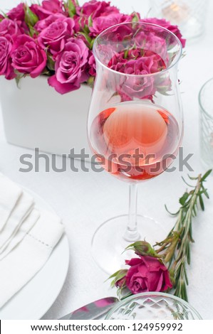 A glass of rose wine and festive dining table setting with pink roses, white tablecloths and napkins, candles, holiday dinner.