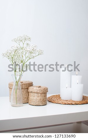 A Branch Of Flowers In A Small Vase, White Candles On The Stand And Two Closed Baskets On A White Table In An Interior